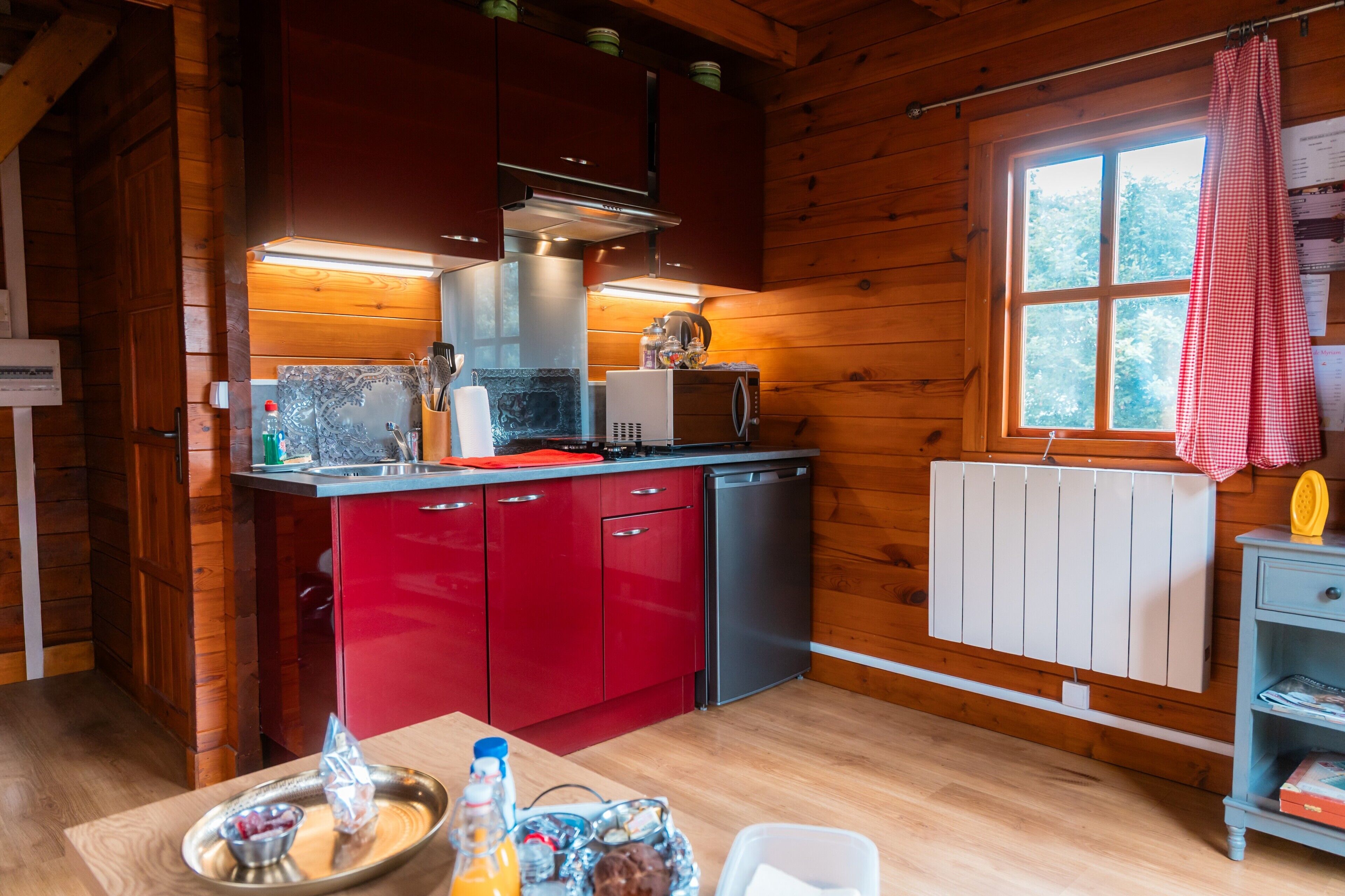 A wooden cabin interior with a kitchen featuring red cabinets, a stainless steel dishwasher, and a window with a red checkered curtain.