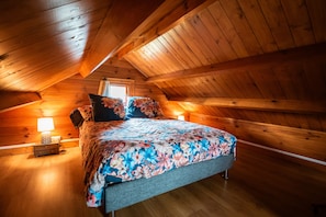 Ein Schlafzimmer mit Holzboden, einem Bett mit Blumenmuster auf der Bettwäsche, einem Nachttisch mit Lampe und einem Fenster.