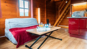 A cozy cabin interior with a wooden floor, a sofa, a dining table with a red tablecloth, and a kitchen area with red cabinets. - Guest Room - Chalet in Pays d'Auge with Garden (Lessard-et-le-Chêne)