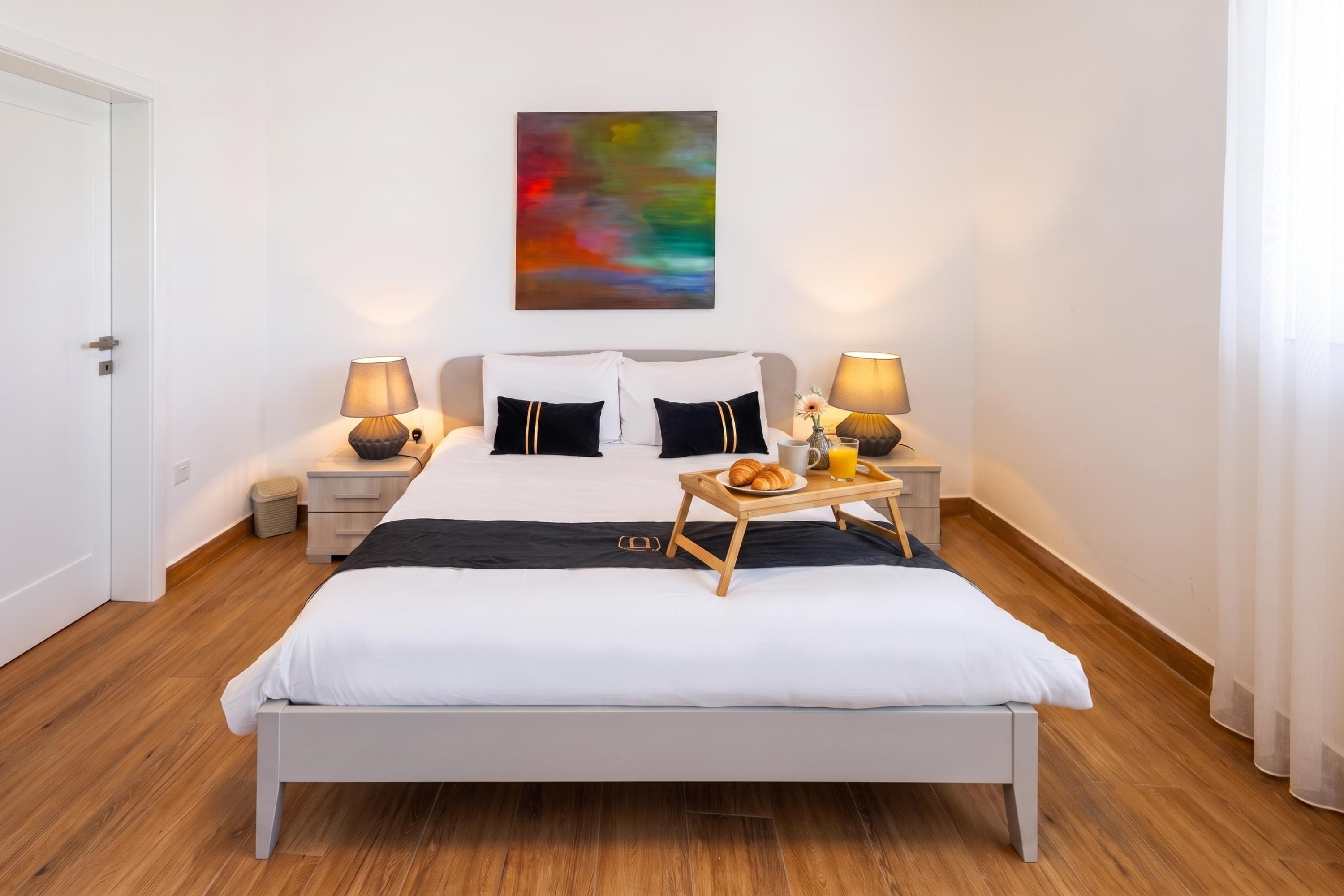A modern bedroom with a large bed, two bedside tables, a bedside lamp, a colorful abstract painting, and a tray with croissants and juice.