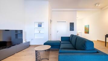 A modern living room with a large flat-screen TV, a blue sectional sofa, a small round coffee table, and a minimalist design.