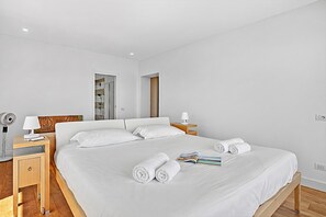 A hotel room with a large bed, two bedside tables, a nightstand, a lamp, a fan, and a wooden floor.