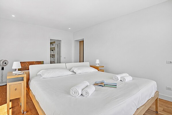 A hotel room with a large bed, two bedside tables, a nightstand, a lamp, a fan, and a wooden floor. - Villa Alessia - 4 bed Villa With Lake Maggiore View (Stresa)