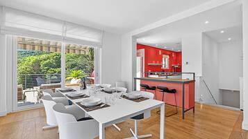 A modern dining area with a white table set for six, a kitchen with red accents, and a view of greenery outside.