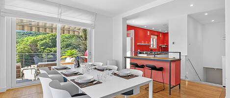 Un comedor moderno con una mesa blanca para seis personas, una cocina con detalles en rojo y vistas al exterior con vegetación.