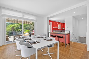 Un comedor moderno con una mesa blanca para seis personas, una cocina con detalles en rojo y vistas al exterior con vegetación.
