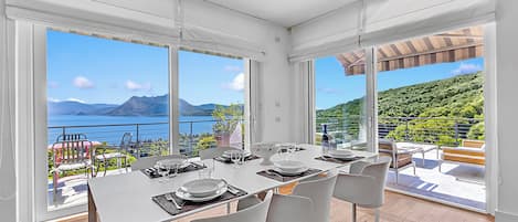 Un coin repas avec une table dressée pour quatre personnes, de grandes fenêtres et une vue sur la mer et les montagnes.