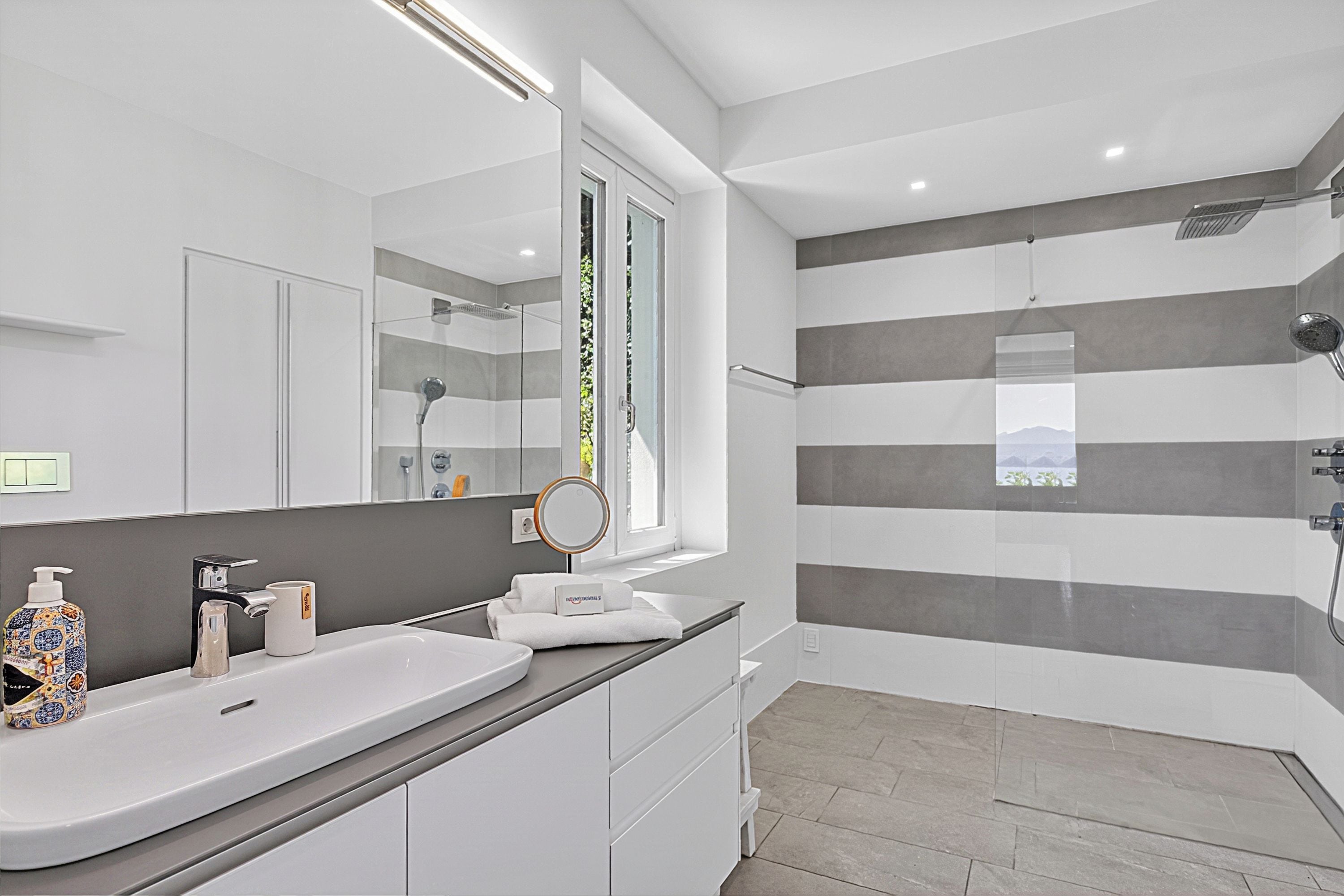 A modern bathroom with a large mirror, a white sink, a shower, and a view of the outdoors.