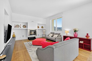 A modern living room with a flat-screen TV, grey sofas, a red ottoman, a fireplace, and a red bookshelf. - Villa Alessia - 4 bed Villa With Lake Maggiore View (Stresa)