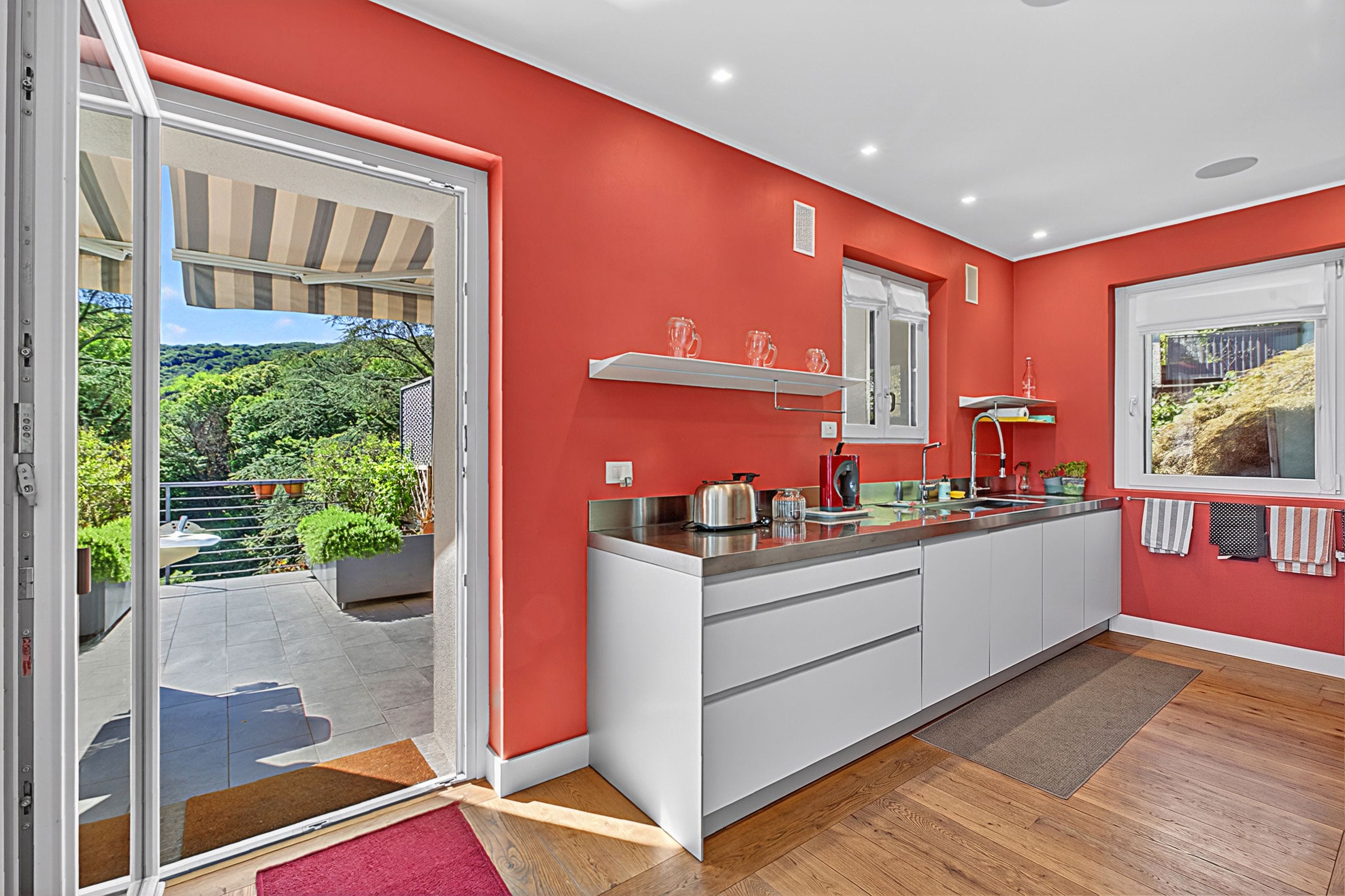 A modern kitchen with a red wall, a sliding glass door leading to a balcony, and a wooden floor.