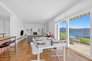 Una cocina moderna con muebles blancos, un comedor con una mesa blanca dispuesta para seis personas, y un amplio ventanal con vistas panorámicas a montañas y un lago.