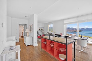 A modern kitchen with a central island, large windows with a view of the sea, and a dining area with white chairs. - Villa Alessia - 4 bed Villa With Lake Maggiore View (Stresa)