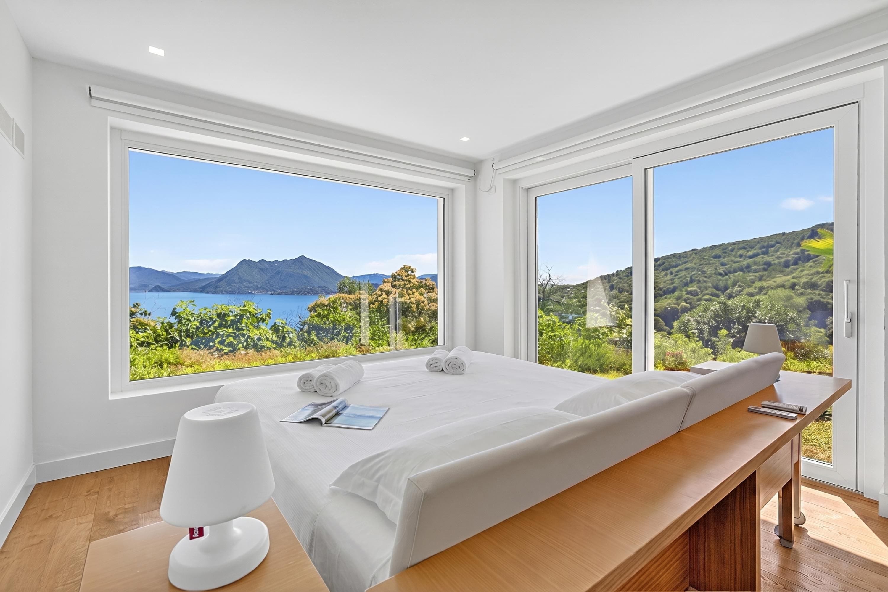 A modern bedroom with a large bed, a bedside table with a lamp, and a view of a mountainous landscape through large windows.