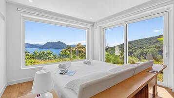 Un dormitorio moderno con una cama grande, una mesita de noche con una lámpara, y vistas a un paisaje montañoso a través de amplias ventanas.
