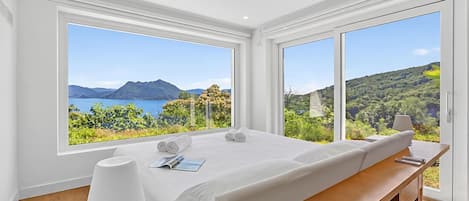 Un dormitorio moderno con una cama grande, una mesita de noche con una lámpara, y vistas a un paisaje montañoso a través de amplias ventanas.