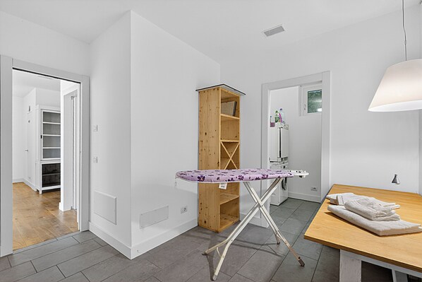 A room with a wooden ironing board, a wooden shelf unit, a table with a folded towel, and a door leading to another room. - Villa Alessia - 4 bed Villa With Lake Maggiore View (Stresa)