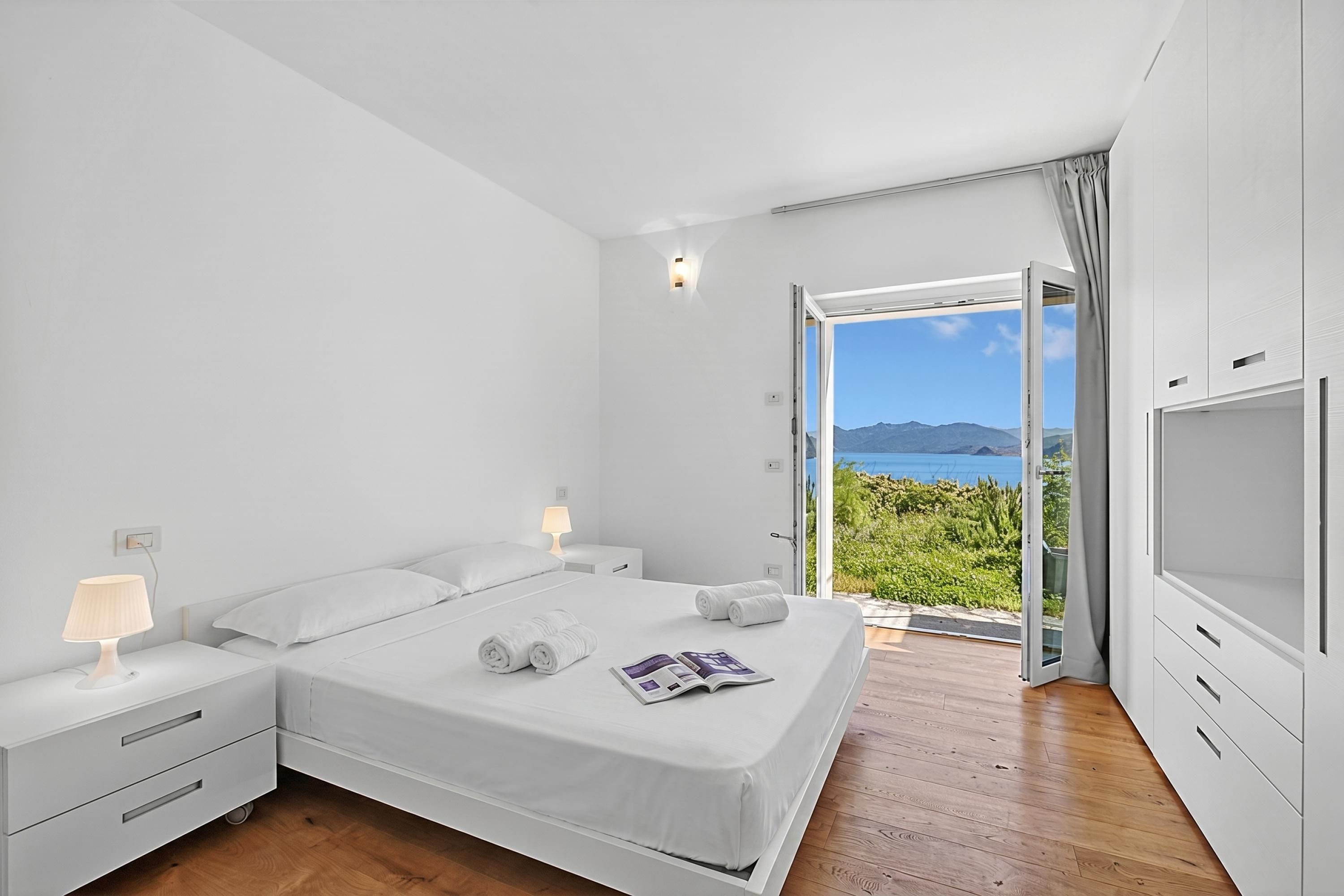 A bedroom with a bed, bedside tables, a lamp, a magazine, and a view of the sea.