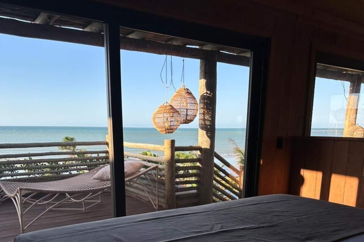 A bedroom with a bed, a view of the ocean, and a balcony with wicker furniture.