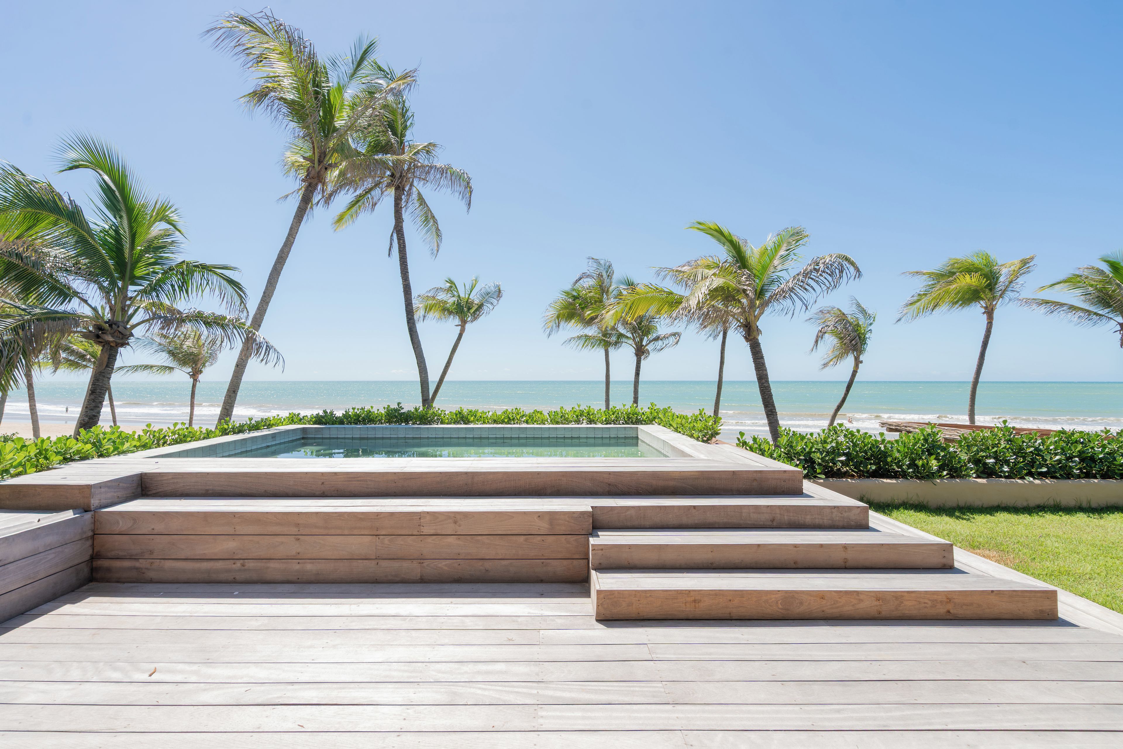 Zona de piscina con escalones que bajan a la piscina, rodeada de palmeras y vistas a la playa.