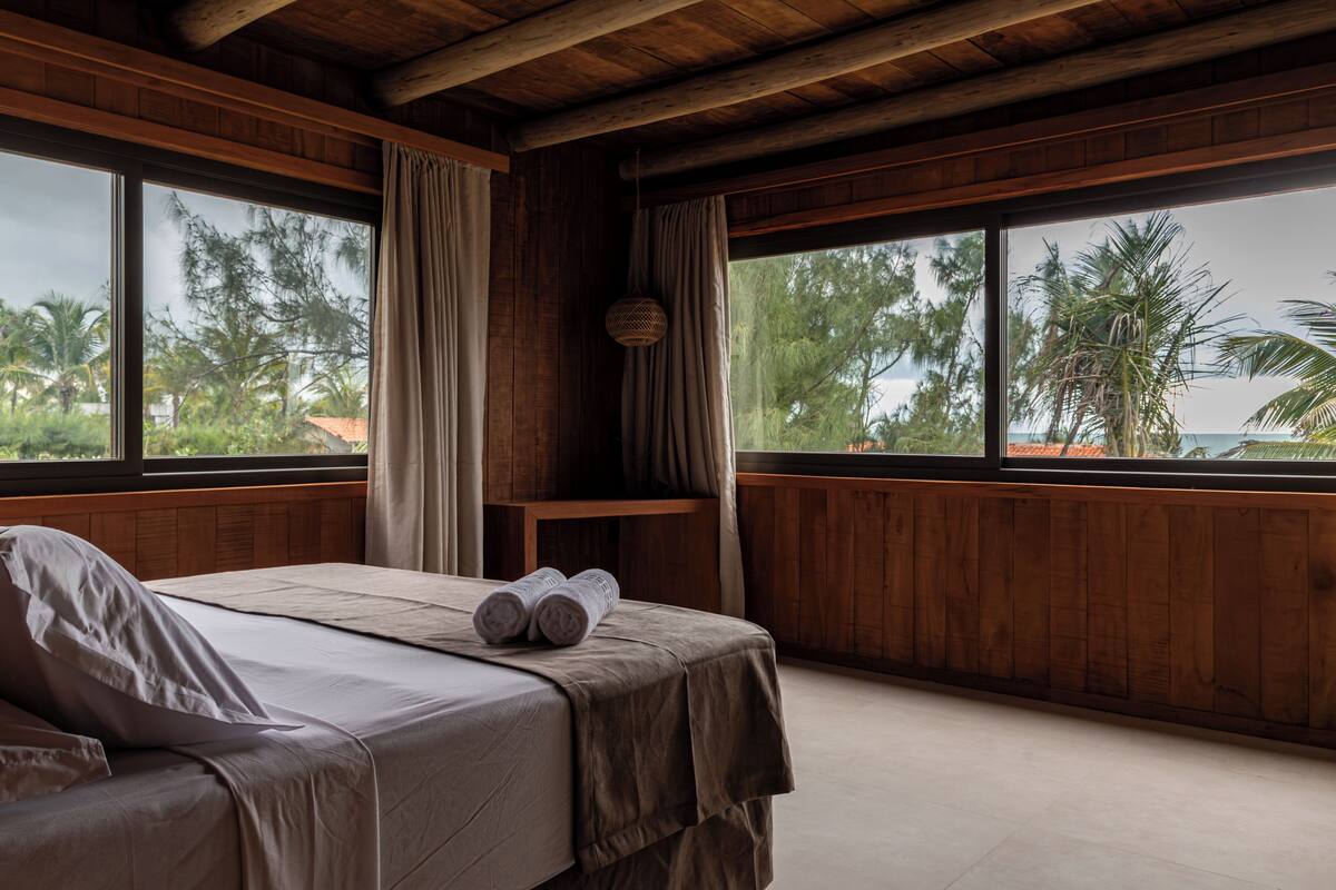 A room with a bed, wooden walls, large windows, and a view of palm trees.