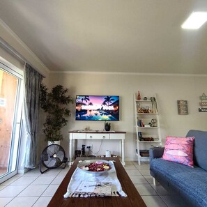 Una sala de estar con televisor, sofá, mesa de centro con flores, ventilador y estantería.