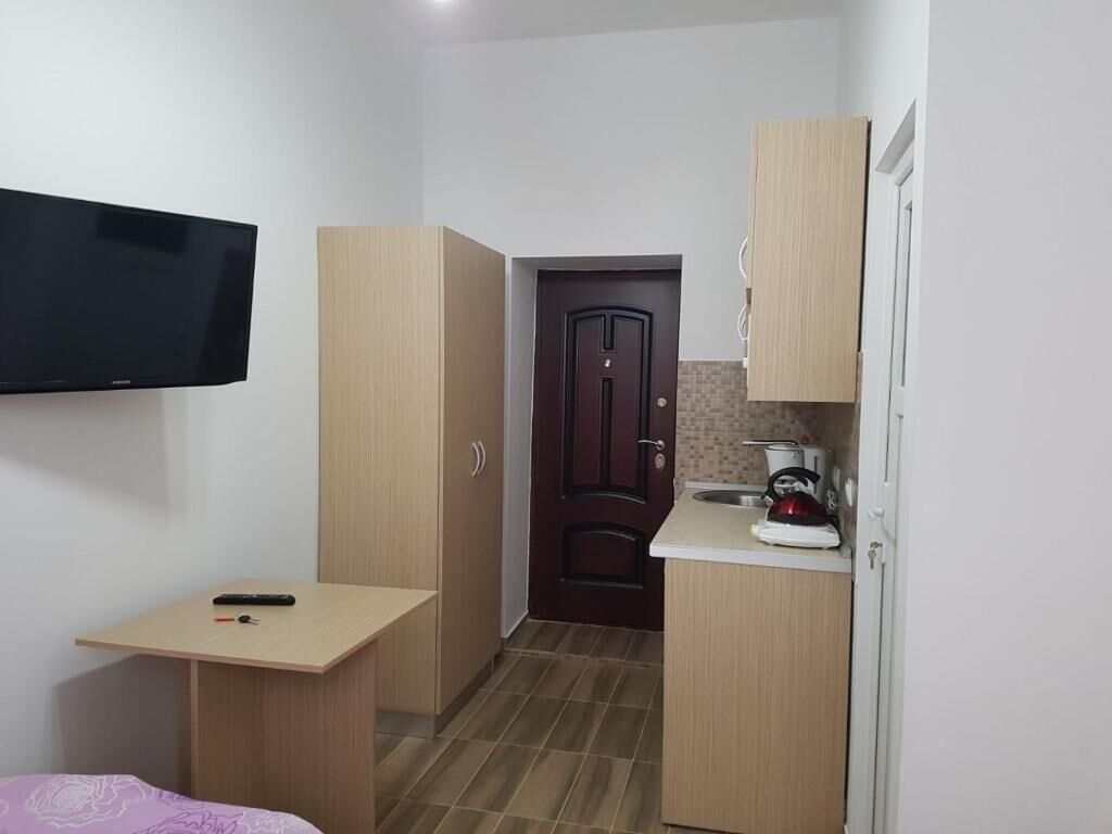 A compact room with a bed, a small table, a TV mounted on the wall, a kitchenette with a stove, and a door leading to another room.
