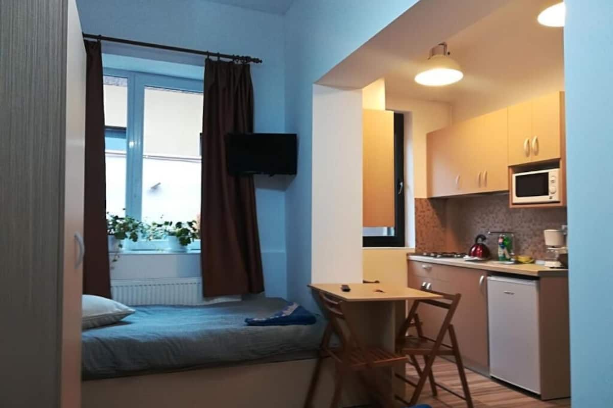 Een compacte kamer met een bed, een klein tafeltje, een stoel, een kitchenette en een aan de muur bevestigde televisie.