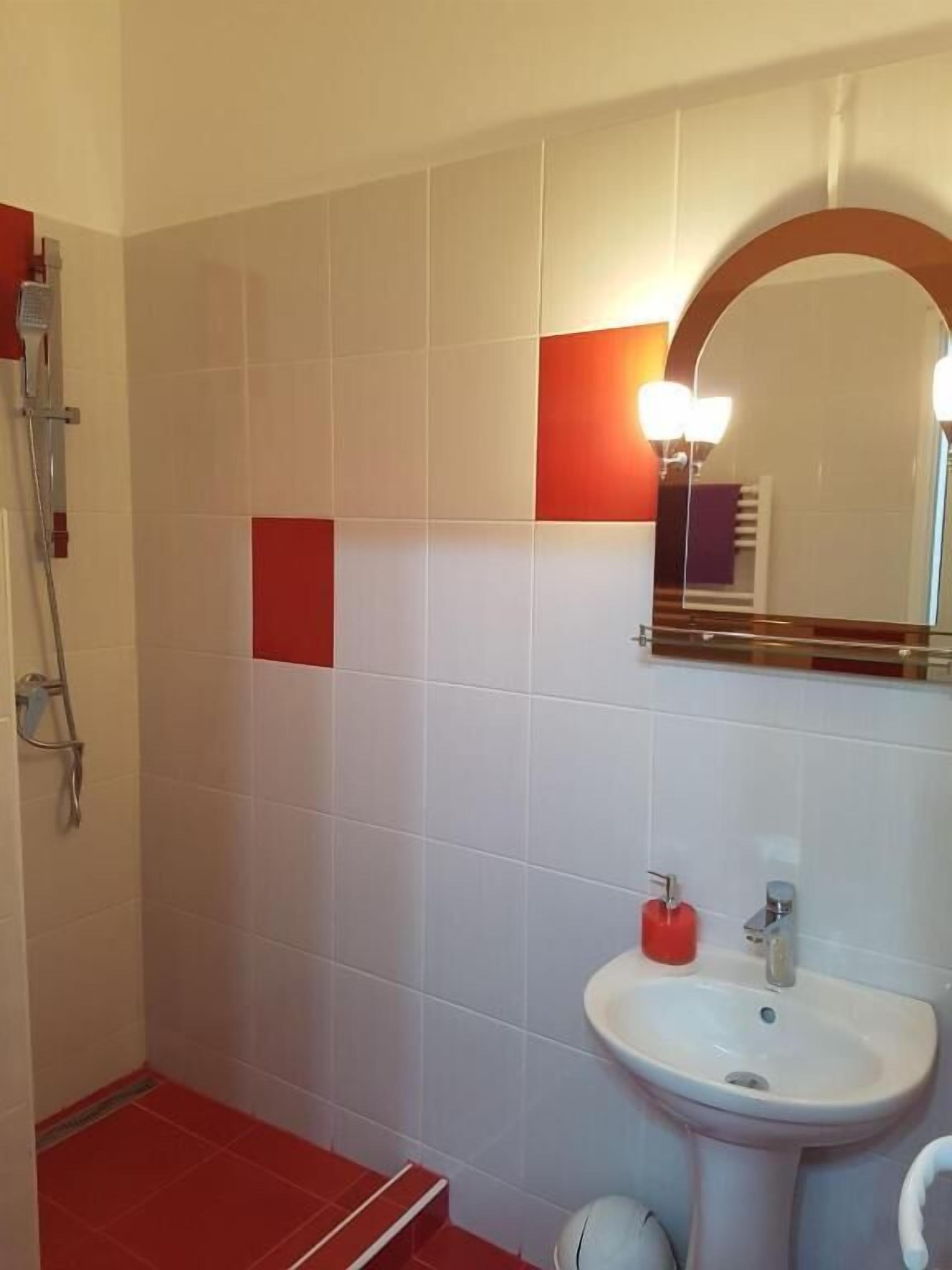 A bathroom with white and red tiles, a sink, a mirror with lights, and a shower area.