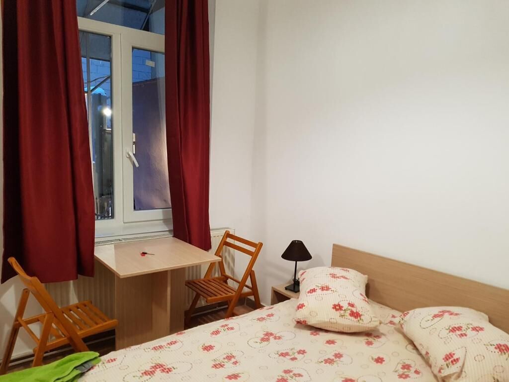 Una habitació petita amb un llit, una cadira de fusta, una taula i una finestra amb cortines.