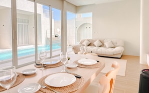 Zona de menjador amb vista a una piscina, un sofà i una taula de dinar preparada amb plats i gots.