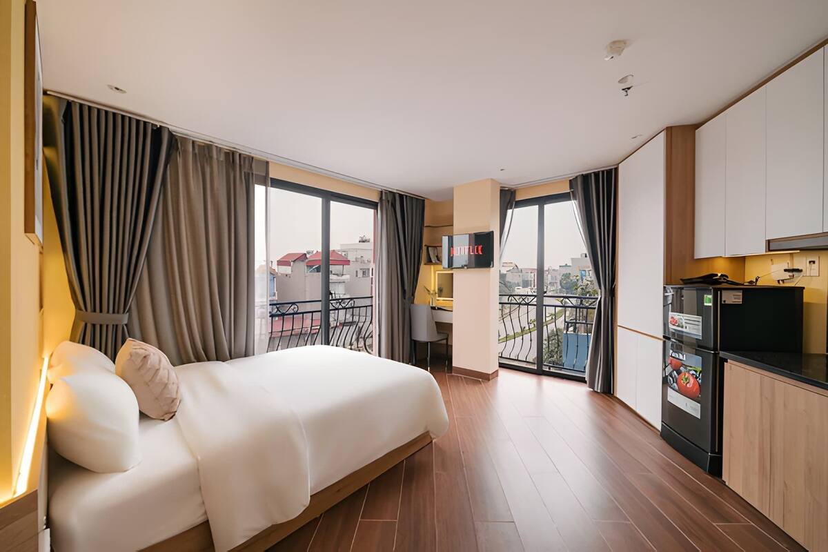 A hotel room with a bed, a kitchenette, a TV, and a balcony with a city view.