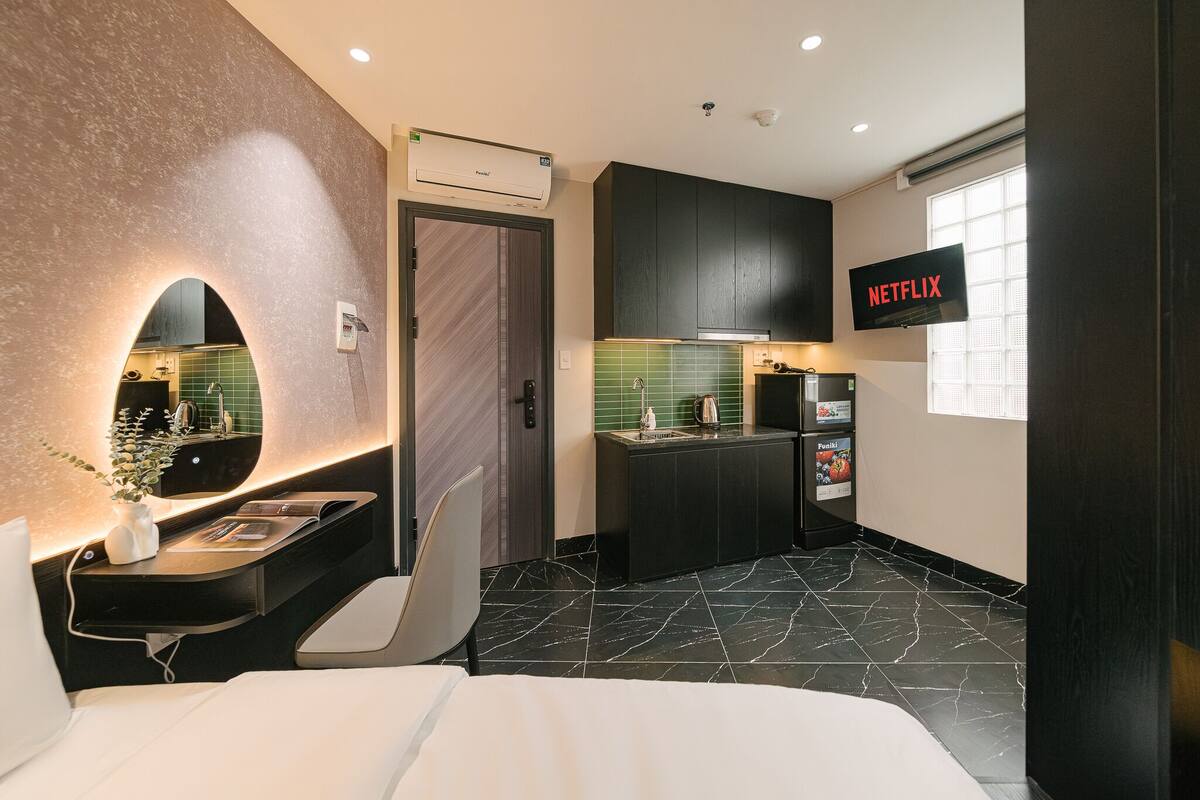 A compact hotel room with a bed, a vanity with a mirror, a small kitchenette, a TV displaying 'NETFLIX,' and a window.