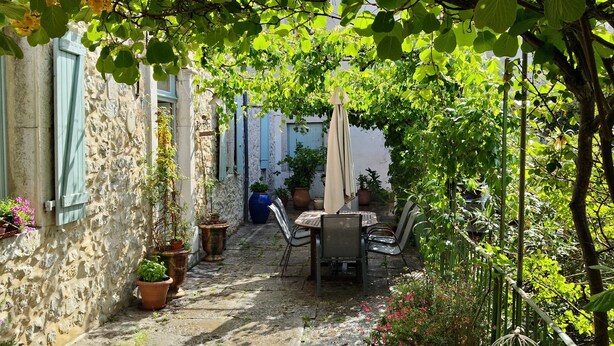Een patio met stenen bestrating, een tafel en stoelen, omgeven door plantenpotten en een pergola met klimplanten.
