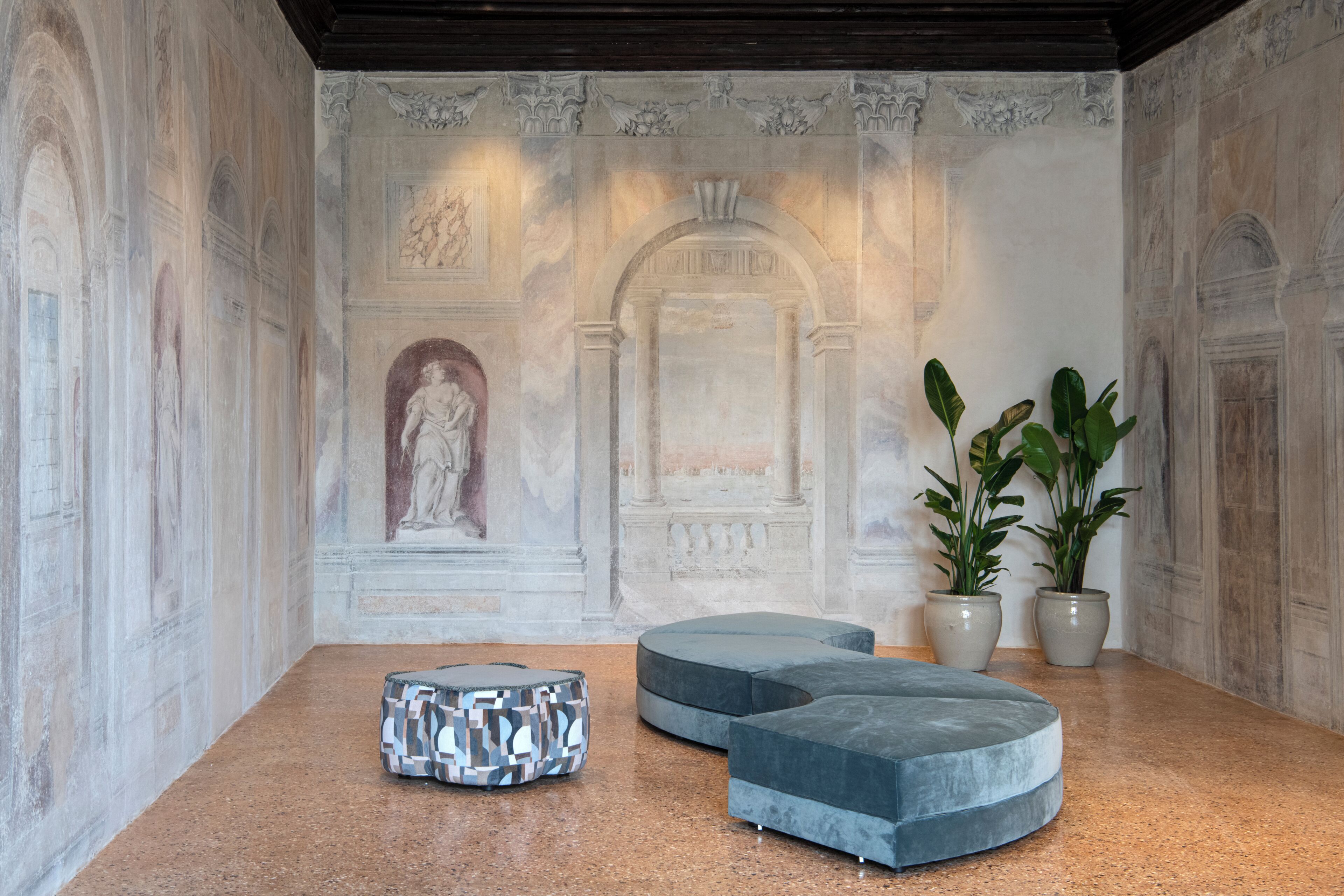 Une pièce dotée d’un sol en marbre, d’une fresque murale de style classique et de deux coins salon avec des fauteuils recouverts d’un tissu gris.