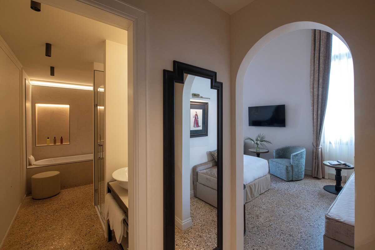 Habitación de hotel con baño, cama, zona de estar y televisor montado en la pared.