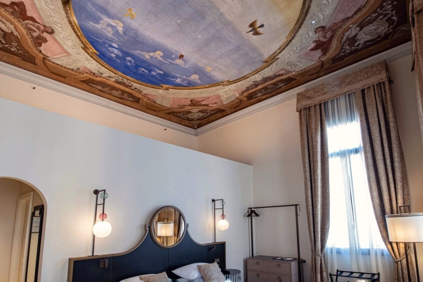 A hotel room with a large bed, two wall-mounted lamps, a ceiling mural, and a window with curtains.