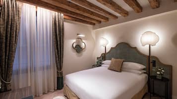 A hotel room with a large bed, wooden ceiling beams, a nightstand with a lamp, and a window with curtains.