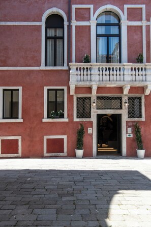A building with a red facade, multiple windows, a balcony with a balustrade, and potted plants. - Palazzo De Polo (Venice)