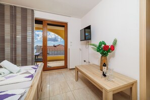 Una habitación de hotel con una cama, una mesa de madera con una botella y vasos, un televisor montado en la pared y vista a un edificio a través de una puerta.