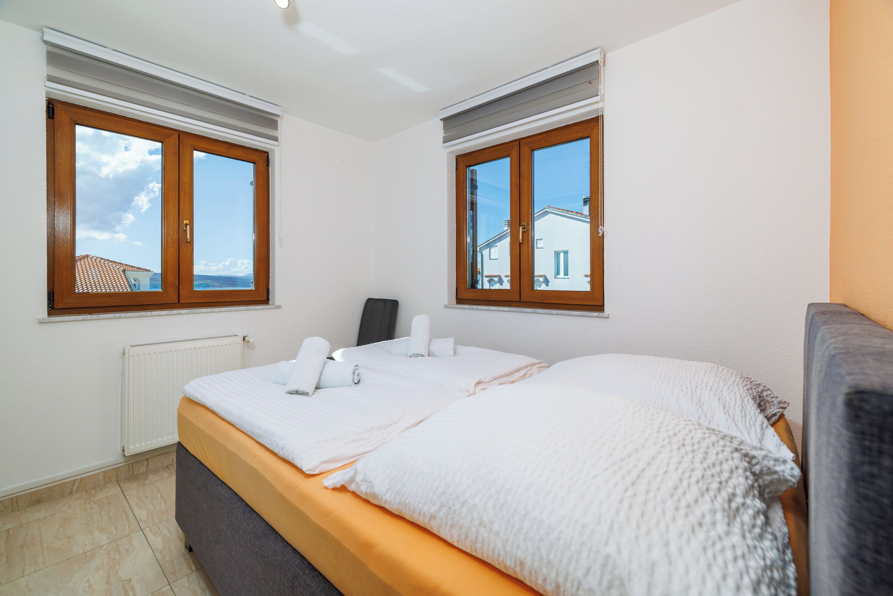 A bedroom with a bed, two windows, a radiator, and a view of the outside.