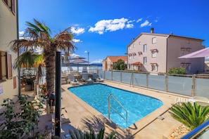 A swimming pool with lounge chairs, a palm tree, and a building with a red-tiled roof. - Cozy vacation apartment with pool and terrace in Crikvenica (Crikvenica)