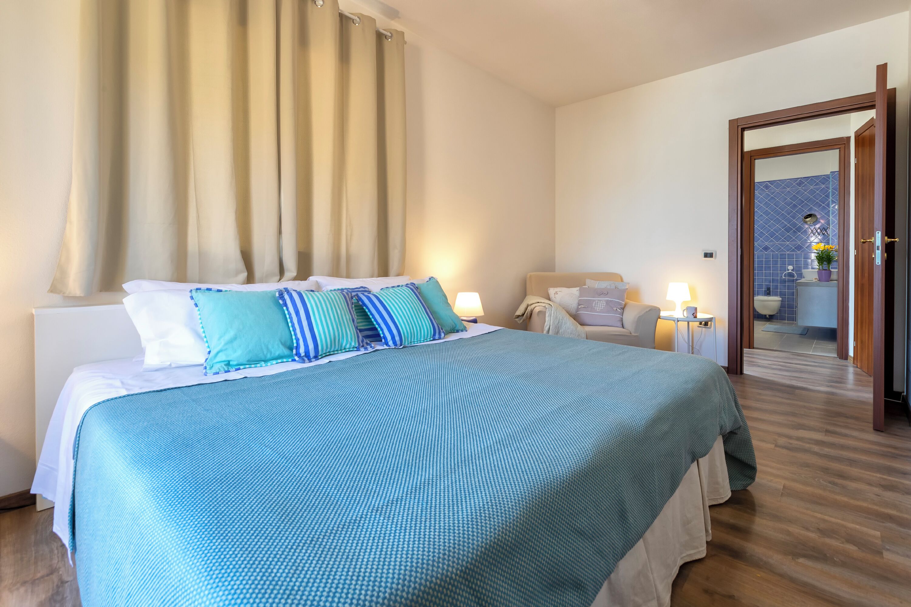 Camera d'albergo con un letto grande, un comodino, un tavolino e un bagno parzialmente visibile attraverso una porta aperta, con pavimento in legno.