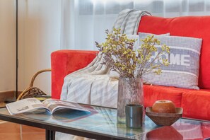 A red sofa with a 'HOME' pillow, a clear glass table with a magazine, a vase with dried flowers, and a wooden bowl. - SARDINIA GUEST HOUSE (Olmedo)