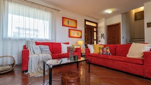 Sala de estar con dos sofás rojos, una mesa de centro de vidrio y un reposapiés marrón. Hay dos cuadros enmarcados en la pared, una puerta y una ventana con cortinas de gasa.
