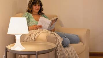 Una persona sentada en un sillón beige leyendo un libro, con una lámpara de mesa blanca a su lado.