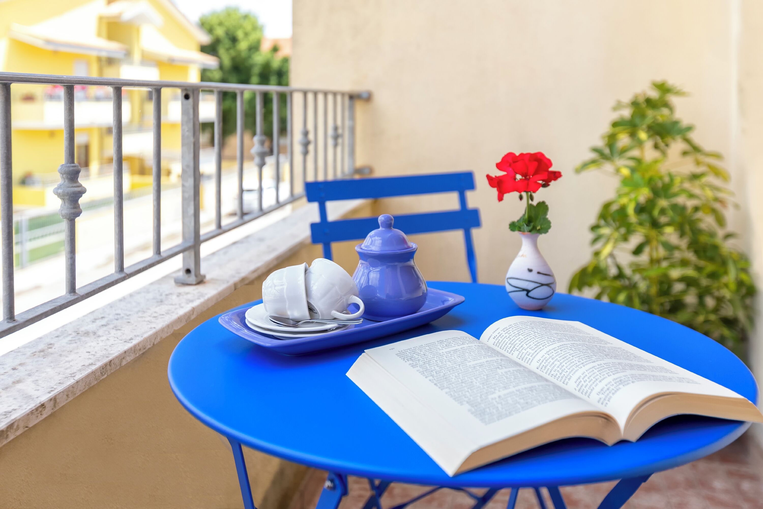 A blue table with a book, teapot, cups, and a vase with flowers on a balcony.