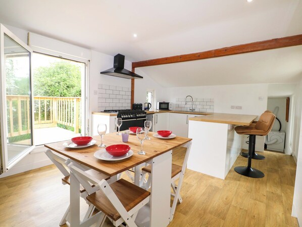 A modern kitchen with a gas stove, microwave, and sink. There is a dining area with a wooden table and white chairs. A balcony is visible with a wooden railing. - Woodside (Battle)
