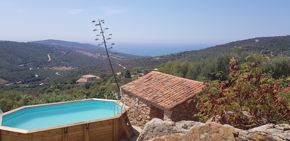 Ein hölzerner Pool mit einer Steinstruktur in der Nähe, umgeben von Grün und Blick auf ferne Hügel.