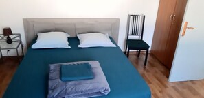 Ein Bett mit zwei Kissen, einer zusammengelegten Decke, einem Stuhl, einem Nachttisch mit Lampe und einem Kleiderschrank.