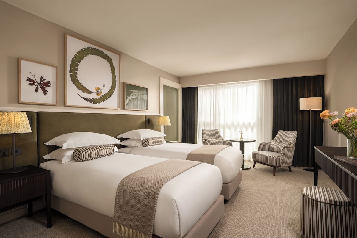 A hotel room with two beds, a desk, a chair, a lamp, and artwork on the wall.
